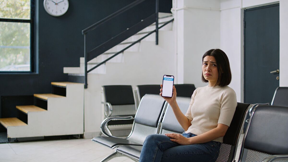 A concerned woman sits in a waiting room holding a smartphone displaying an appointment booking screen for medical services.